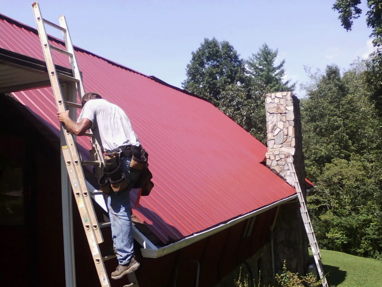 Roofer on a ladder beside a red metal roof and stone chimney — Soffit & Fascia Repair by Roofing Brothers in Ebony